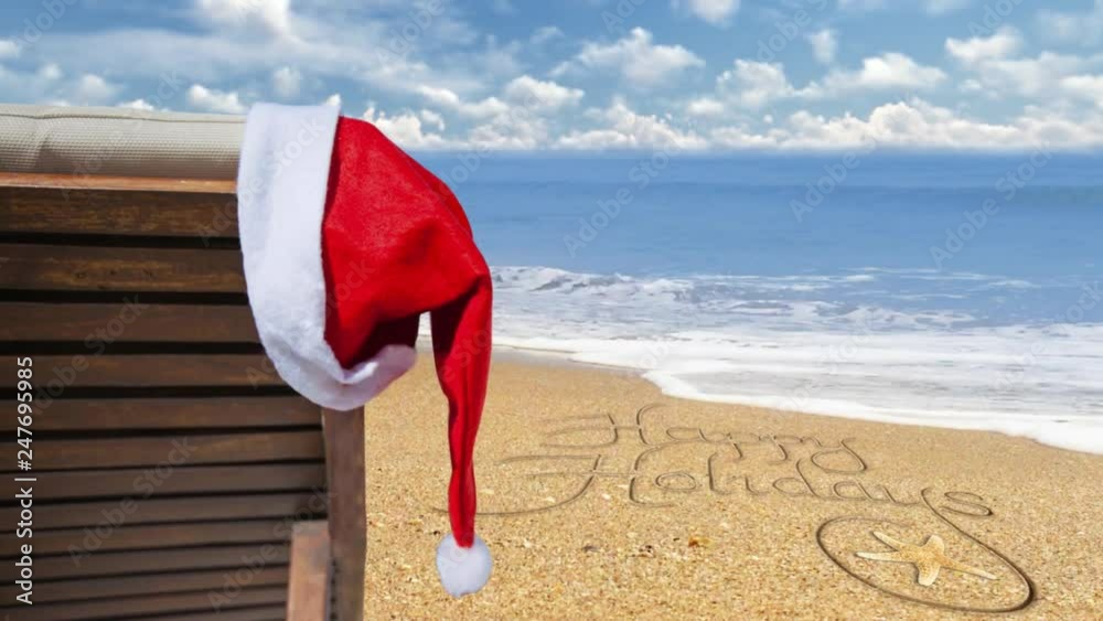 Happy Holidays from the Beach 4K features a beach chair with a Santa hat hanging and gently blowing in the wind with an ocean in the back ground and Happy Holidays written in the sand