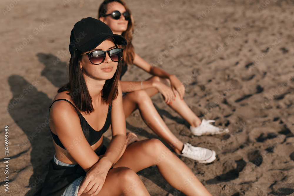 Dark-haired stylish cute woman with short hairstyle wearing cap and glasses sitting with a friend on sandy beach and enjoys sunset on the ocean