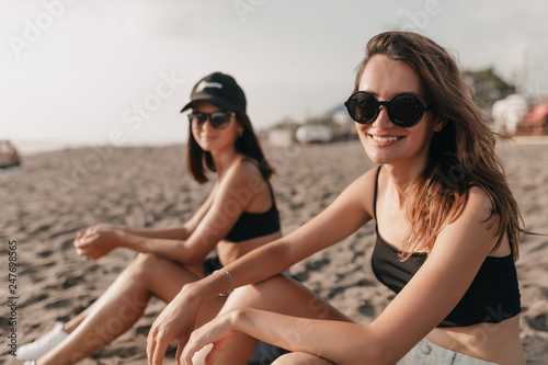 Charming lovely smiling two ladies in black swim suits and glasses sitting on the beach in sunlight posing at camera and enjoyng life on the beach © PhotoBook