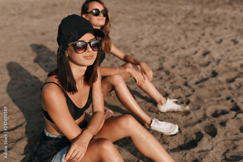 Obraz na płótnie Dark-haired stylish cute woman with short hairstyle wearing cap and glasses sitting with a friend on sandy beach and enjoys sunset on the ocean w salonie