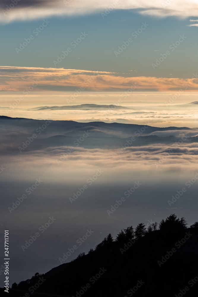 Beautiful view of Umbria valley (Italy) covered by a sea of fog at sunset, with beautiful warm colors and trees silhouettes in the foreground
