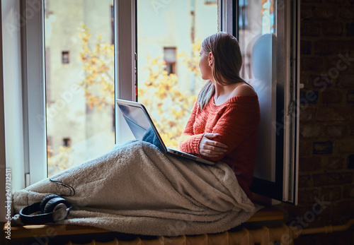 A young woman with glasses and a warm sweater covered her legs with a blanket, with a laptop sitting on the windowsill