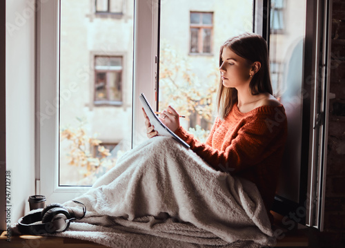 Beautiful girl in a warm sweater covered her legs with a blanket, makes notes in her notebook sitting on the window sill next to the open window