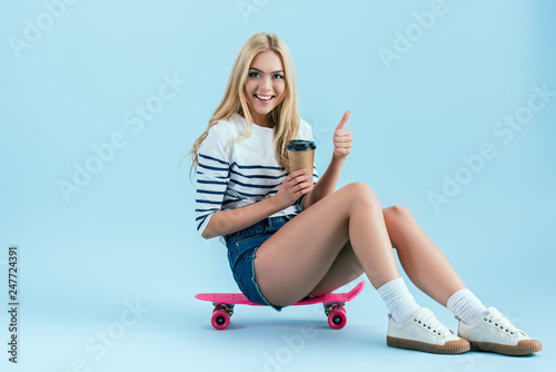 Cheerful girl with cup of coffee sitting on skateboard and showing thumb up on blue background