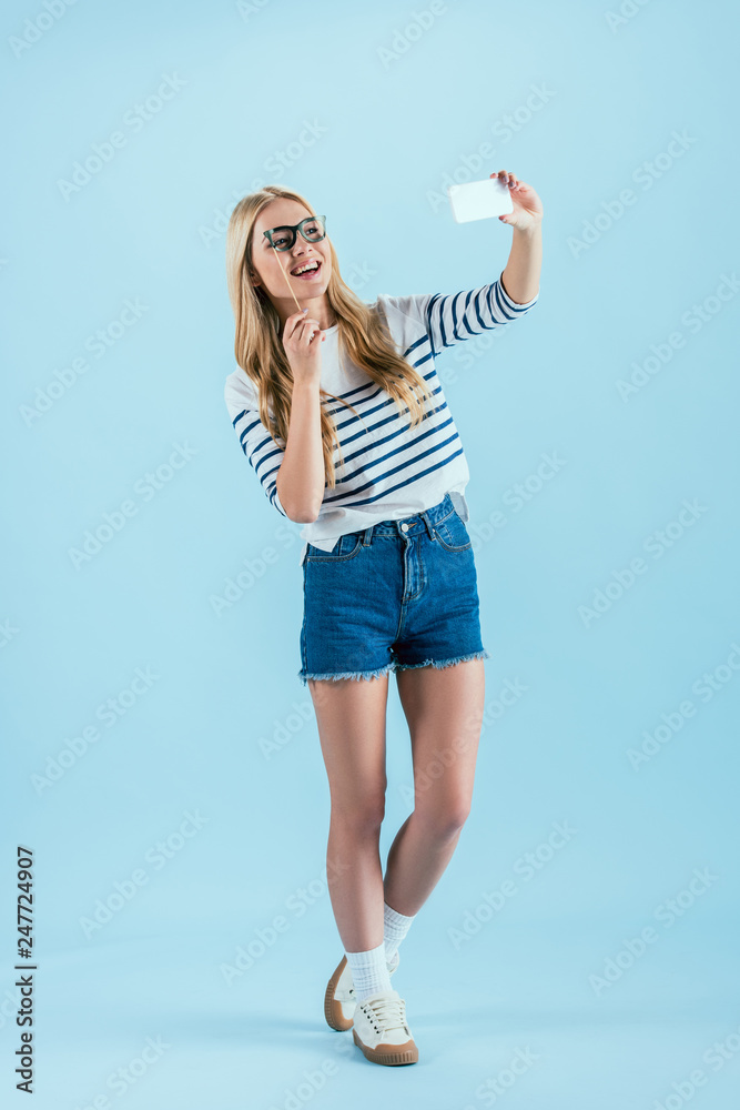Full length view of laughing woman making selfie with toy glasses on blue background Full length view of laughing woman making selfie with toy glasses on blue background