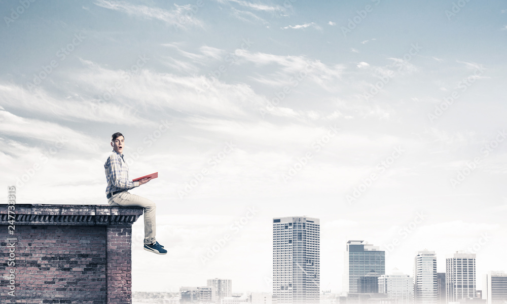Man on roof edge reading book and cityscape at background Man on roof edge reading book and cityscape at background