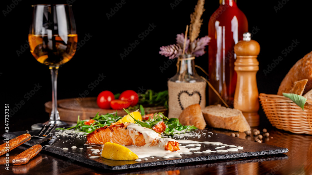 The concept of Italian cuisine. Salmon fillet with arugula, cherry tomatoes and parmesan cheese in creamy lemon sauce. A glass of white wine on the table. Dishes in the restaurant.