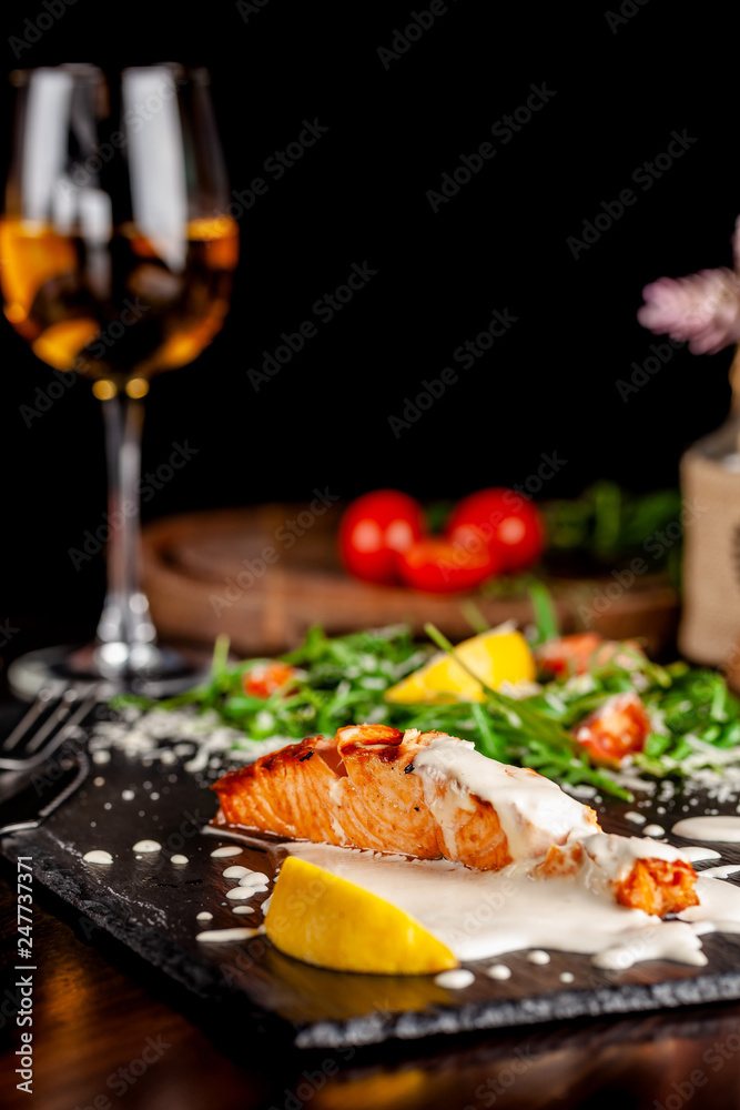 The concept of Italian cuisine. Salmon fillet with arugula, cherry tomatoes and parmesan cheese in creamy lemon sauce. A glass of white wine on the table. Dishes in the restaurant. The concept of Italian cuisine. Salmon fillet with arugula, cherry tomatoes and parmesan cheese in creamy lemon sauce. A glass of white wine on the table. Dishes in the restaurant.