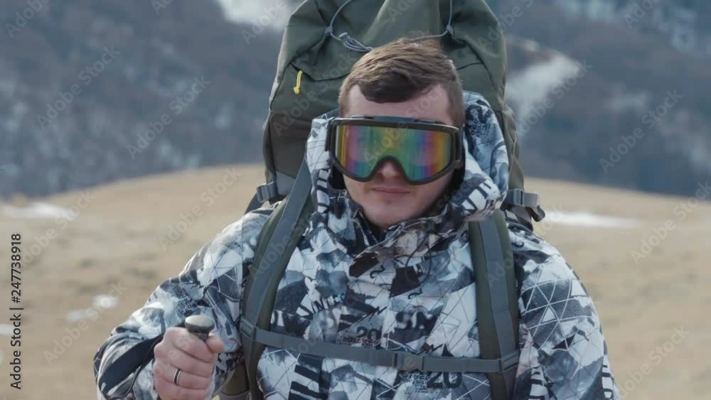 A tourist with trekking poles and a huge backpack against a background of snow-capped mountains in the spring climbs to a top and stops for a halt to drink tea from a thermos. 4K slowmotion. A tourist with trekking poles and a huge backpack against a background of snow-capped mountains in the spring climbs to a top and stops for a halt to drink tea from a thermos. 4K slowmotion.