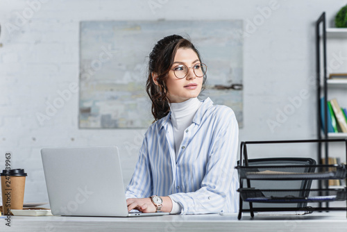 Fotografie Attractive businesswoman in glasses using laptop and looking away
