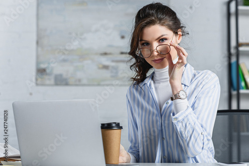 Beautiful businesswoman in glasses holding paper cup and looking at camera at office