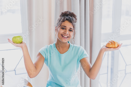 Принти на полотні Beautiful young slender brunette with green apple in one hand and croissant in the other hand on background of vegetables on kitchen table making a difficult choice between healthy and harmful food
