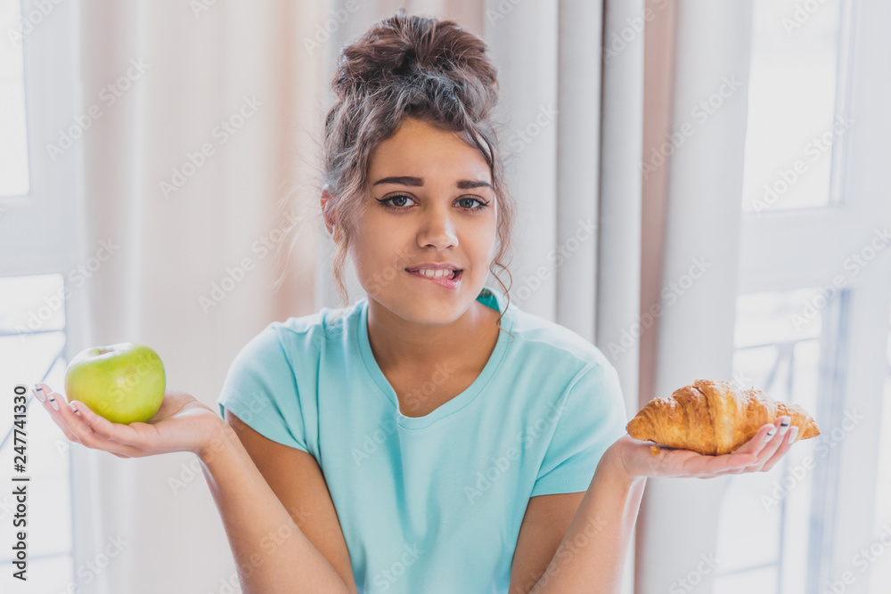 Beautiful young slender brunette with green apple in one hand and croissant in the other hand on background of vegetables on kitchen table making a difficult choice between healthy and harmful food. Beautiful young slender brunette with green apple in one hand and croissant in the other hand on background of vegetables on kitchen table making a difficult choice between healthy and harmful food.