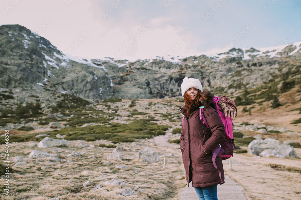 Obraz premium Brunette girl with a hat on her head and a travel backpack contemplates the landscape that surrounds her from a valley next to the mountains.