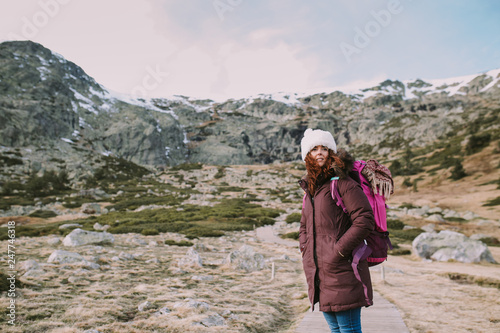 Brunette girl with a hat on her head and a travel backpack contemplates the landscape that surrounds her from a valley next to the mountains.