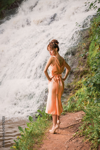 The beautiful young lady in dress standing near the waterfall