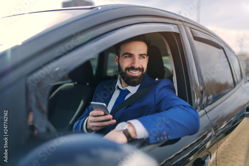 Close up of smiling businessman using smart phone while sitting in his car and leaning on the window.