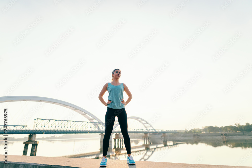 Sporty Caucasian woman standing on the kay with hands on hips in the morning. In background bridge. Full length.