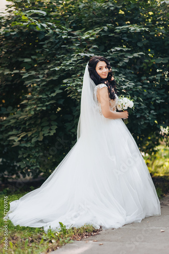 A beautiful bride in a white dress is standing with a bouquet in the park with greenery. Wedding portrait of pretty brunette in the green garden.