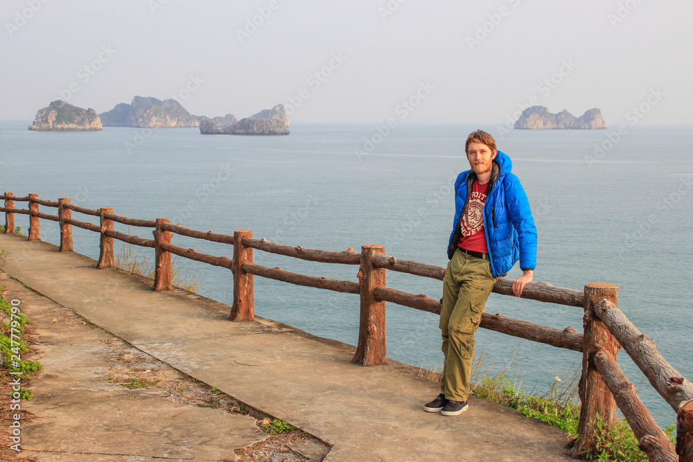 A young white attractive male blond with a beard in a blue down jacket stands against the backdrop of a cliff and the sea in Ha Long Bay, Vientam A young white attractive male blond with a beard in a blue down jacket stands against the backdrop of a cliff and the sea in Ha Long Bay, Vientam