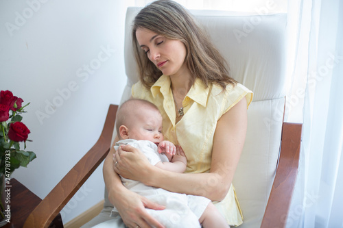 Young mother holding her baby, rocking him to sleep, sitting in armchair photo