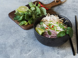Traditional Vietnamese soup- pho ga in black bowl with chicken and rice noodles, mint and cilantro, green and red onion, chili, bean sprouts and lime on grey background. Asian food.