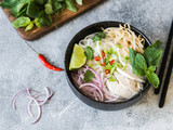 Traditional Vietnamese soup- pho ga in black bowl with chicken and rice noodles, mint and cilantro, green and red onion, chili, bean sprouts and lime on grey background. Asian food.