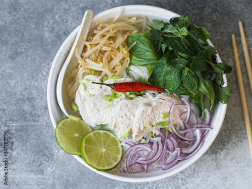 Traditional Vietnamese soup- pho ga in white bowl with chicken and rice noodles, mint and cilantro, green and red onion, chili, bean sprouts and lime on grey background. Asian food. Copy space