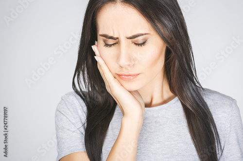 Young European woman isolated on white background suffering from severe toothache, feeling pain so strong that she is pressing fingers to cheek to calm it down, looking desperate.