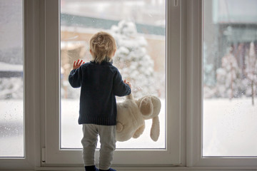 Toddler child standing in front of a big french doors, leaning against it looking outside at a snowy nature .