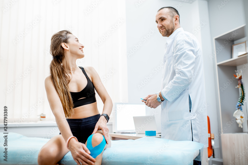 Fototapeta premium Young woman patient sitting with kinesio tape on her knee during the medical treatment with therapist in the office