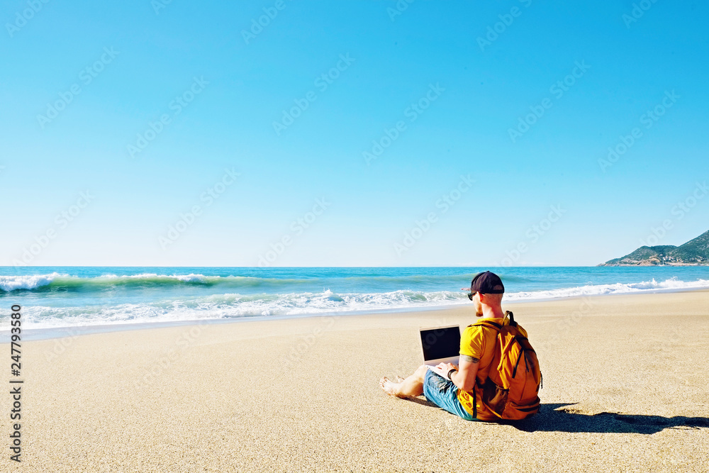 Traveler writing blog entry on white laptop, sitting at exotic empty beach. Freelance remote work concept. Self employed fit young male in bright yellow t-shirt coding. Copy space, sea view background