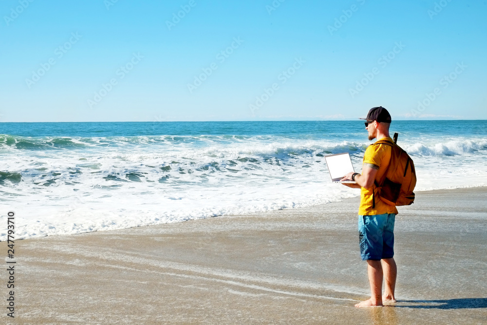 Fit travel blogger holding laptop, writing article on white laptop, standing on empty beach. Freelance remote work concept. Self employed man in yellow t-shirt coding. Copy space, sea view background.