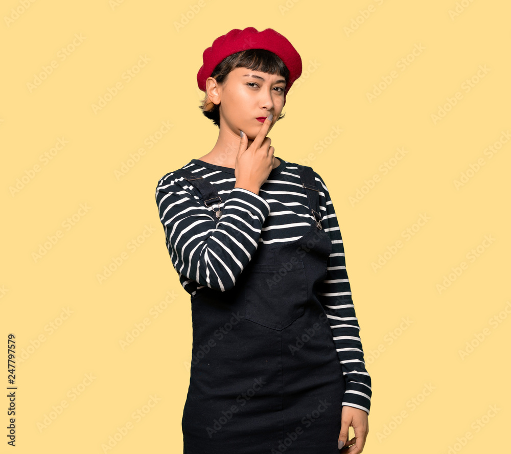 Young woman with beret covering mouth with hands for saying something inappropriate over yellow background