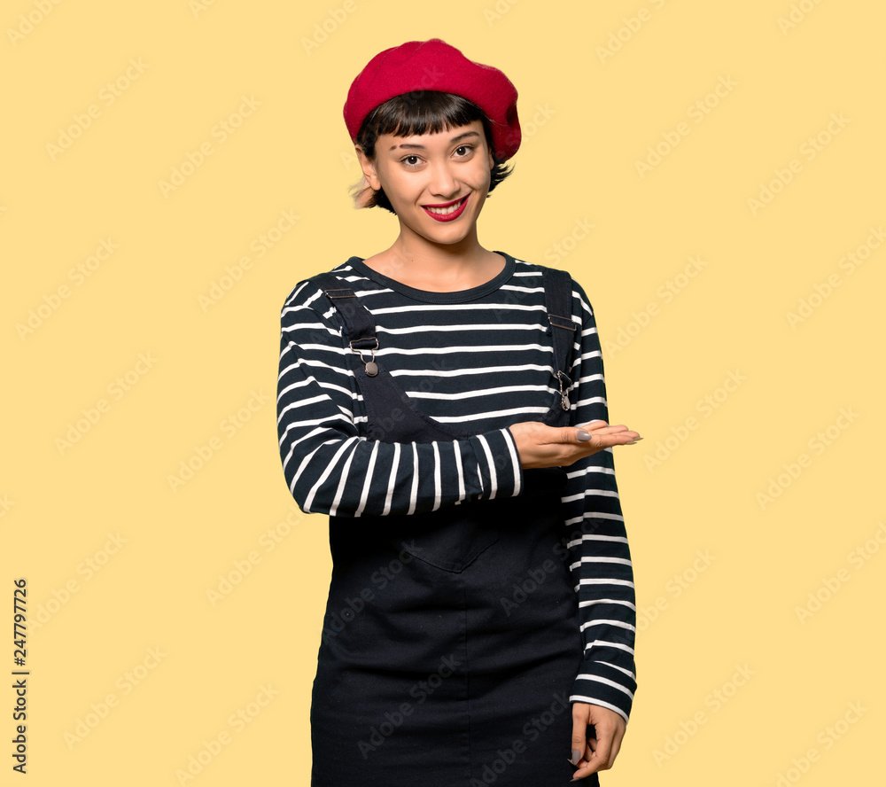 Young woman with beret presenting an idea while looking smiling towards over yellow background Young woman with beret presenting an idea while looking smiling towards over yellow background