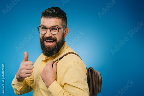 cheerful bearded man with backpack showing thumb up, isolated on blue