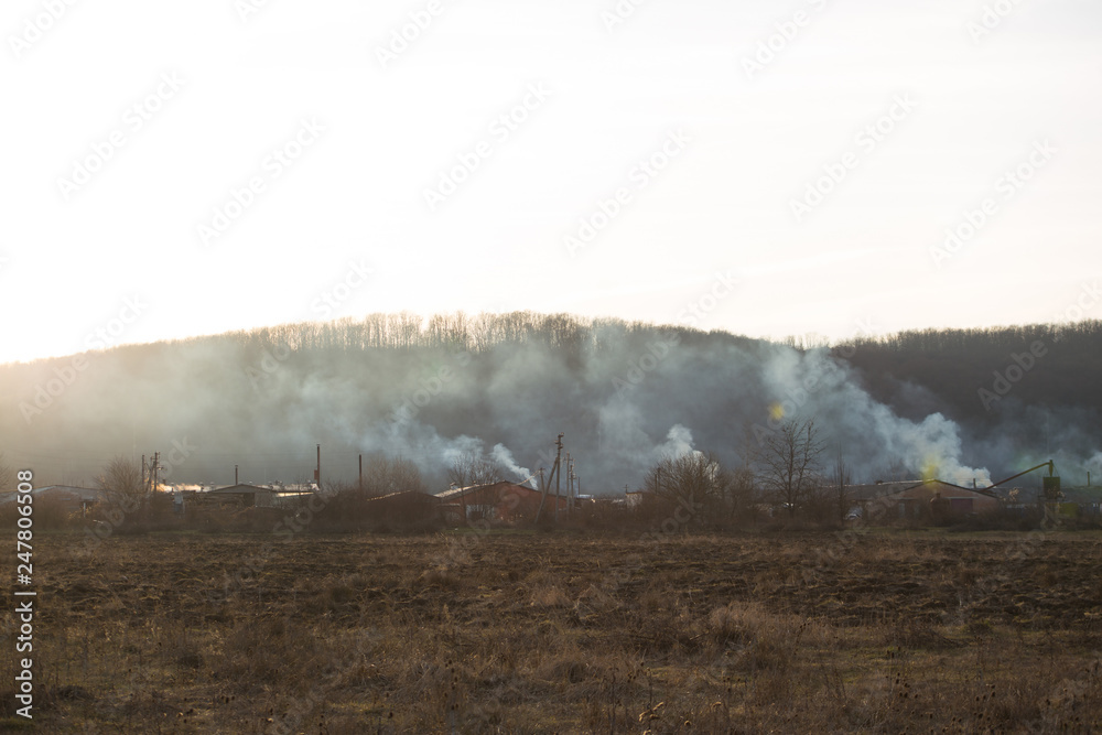 Burning grass near forest with lots of smoke next to wooden houses at sunset against mountain and bright sun. Forest fire. Burning grass near forest with lots of smoke next to wooden houses at sunset against mountain and bright sun. Forest fire.