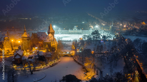 Budapest, Hungary - Aerial view of the beautiful snowy Vajdahunyad Castle in City Park at blue hour with Christmas market, ice rink and Heroes' Square at background during snowing