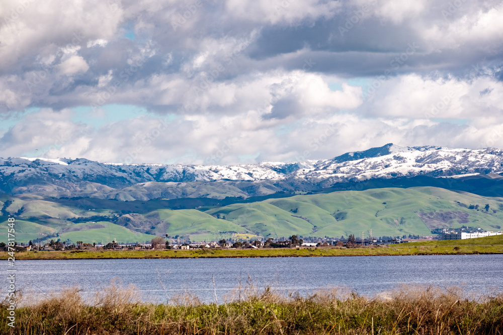 Panoramic view towards green hills and snowy mountains on a cold winter day taken from the shores of a pond in south San Francisco bay area; San Jose, California