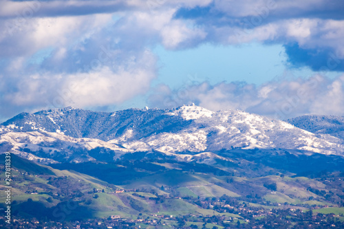 View towards Mt Hamilton and the Lick Observatory building on a sunny winter day; green hills in the foreground and snow covered peaks in the background; San Jose, south San Francisco bay, California