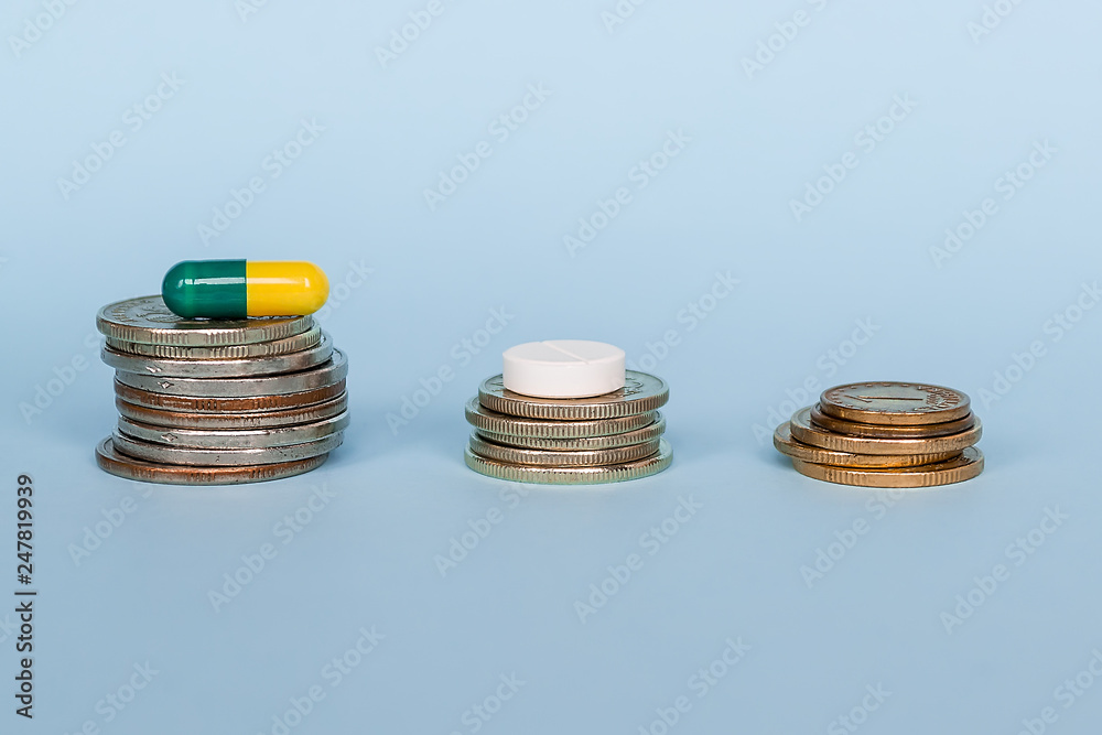 A medicine capsule is on a stack of coins on a blue background close-up. The concept of the high cost of medical drugs A medicine capsule is on a stack of coins on a blue background close-up. The concept of the high cost of medical drugs