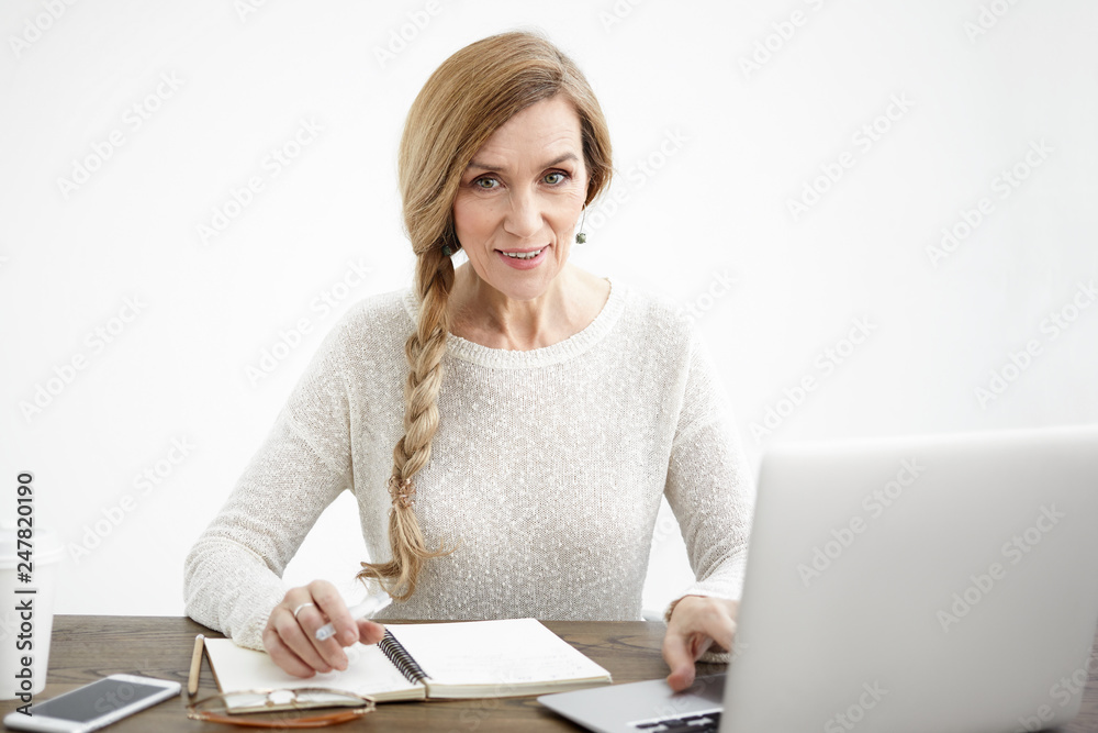 Portrait of attractive confident mature female on retirement enjoying her work, writing down ideas, thoughts and plans in her copybook, using wireless internet connection on laptop computer Portrait of attractive confident mature female on retirement enjoying her work, writing down ideas, thoughts and plans in her copybook, using wireless internet connection on laptop computer