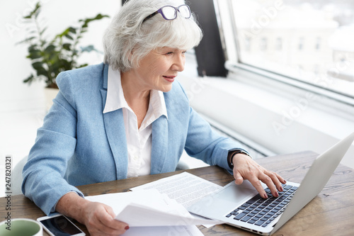 Reputable 65 year old female executive in stylish bkue suit enjoying wireless high speed internet connection while using laptop, analyzing accounts, holding papers in her hand, looking at screen