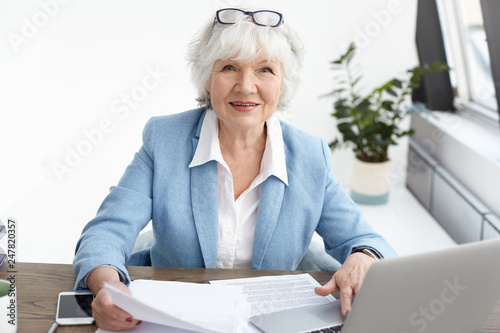 Picture of attractive confident elderly mature female financial adviser with short gray hair looking at camera with smile, studying piece of paper in her hands while working at her office desk