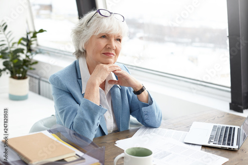 Succesfful skilled attractive elderly female editor of popular fashion magazine sitting at her workplace with papers, mug and open portable computer, clasping hands, having pensive facial expression