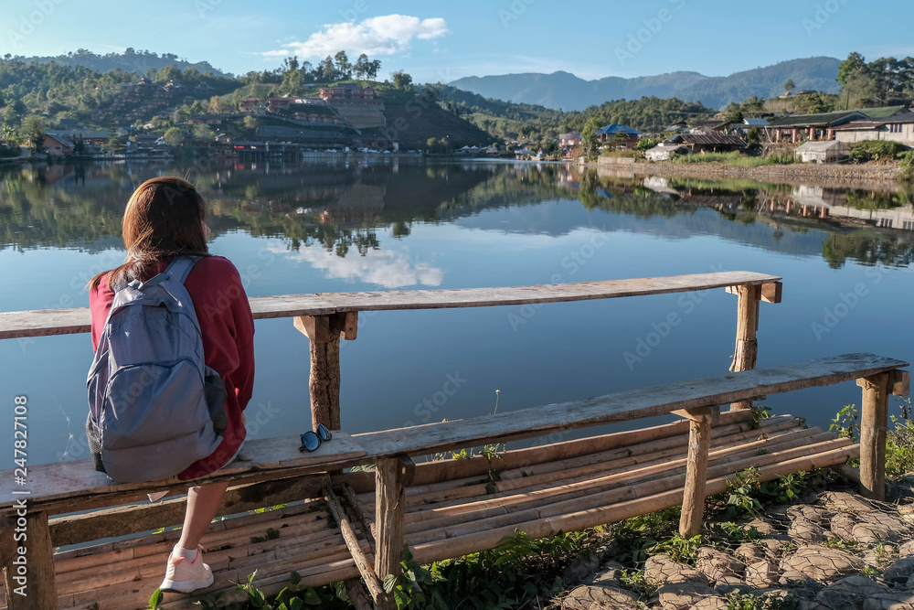 asia woman traveler sit on wooden tabel to  rest and relax looking out at reflection view of lake with chinease style house at ban ruck thai