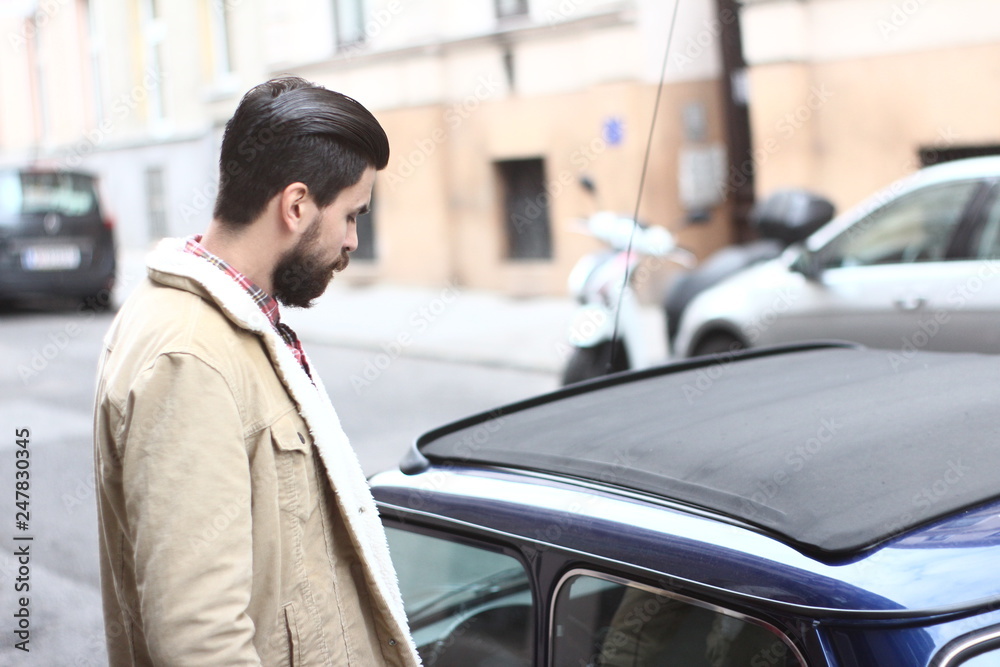 The photo of a face of a handsom bearded man gay in brown jacket on the street outside near a blue car