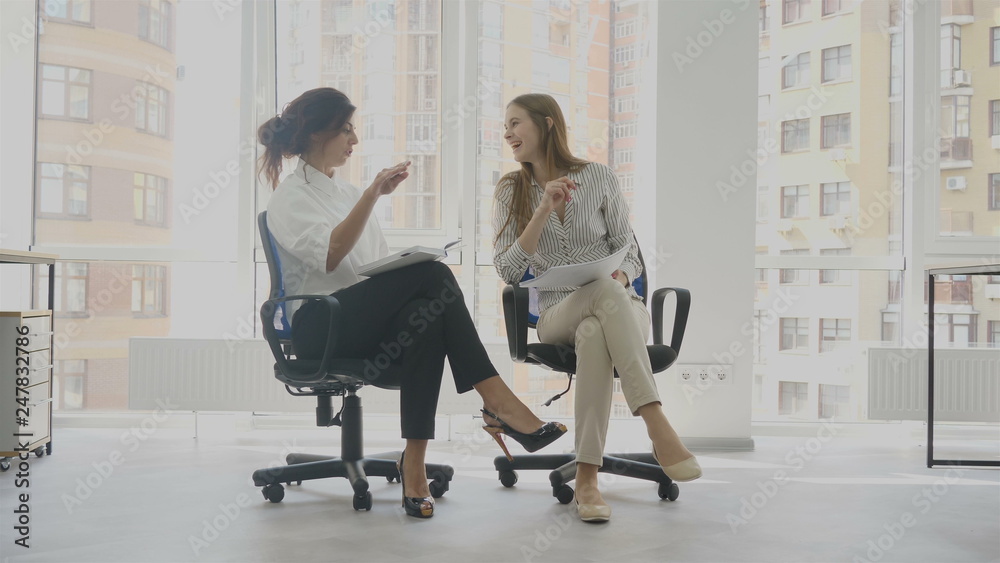 Obraz premium Office workers, two women sitting on chairs talking, one of the women tells a funny story the other laughs