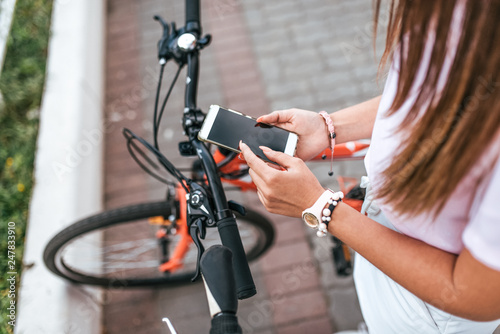 Fototapeta Naklejka Na Ścianę i Meble -  A girl stands with a bicycle in the summer in city. In the hands of a smartphone, writes a message through the application in social networks. Concept search route or map in the city.
