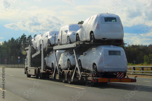 Heavy car carrier truck transports new cars in protective covers on semi-trailer on suburban asphalt highway road on summer day against forest and blue sky - logistics business in Europe, delivery